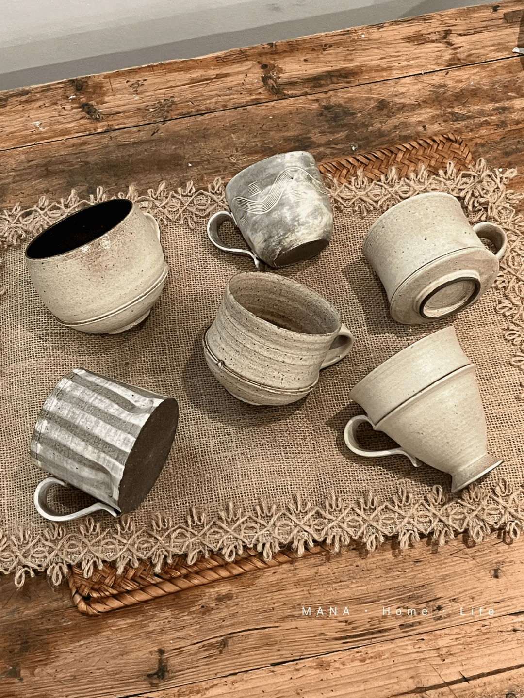 A collection of unique, hand-thrown raw stoneware coffee mugs on a rustic burlap runner showing shape variations.