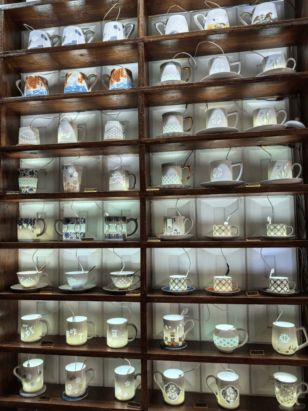 A wooden display shelf filled with illuminated translucent openwork porcelain cups, highlighting the diversity of Jingdezhen ceramics.