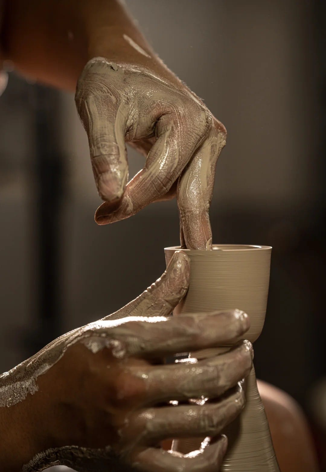 Close-up of a potter's clay-covered hands shaping a ceramic vessel on a pottery wheel in a Jingdezhen workshop.